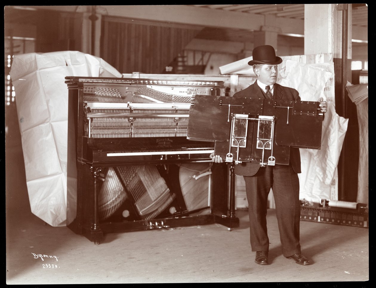 Et piano av Ricca and Son Piano Co. med en mann ved siden av som holder opp en del av pianoverket, 1907 (sølv gelatintrykk) av Byron Company