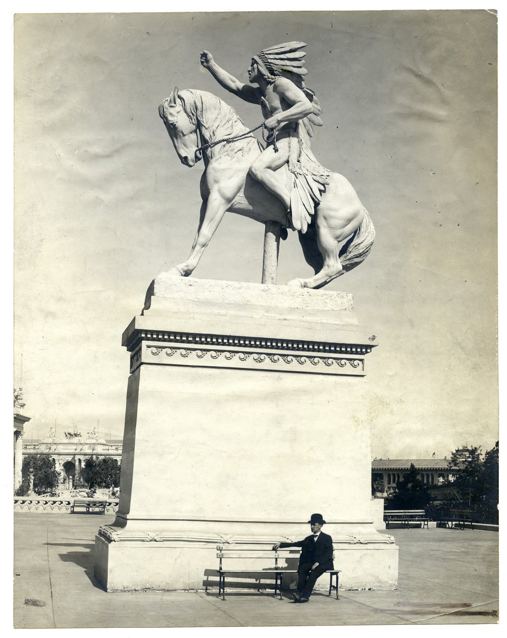 Vertikalt sepia -fotografi som viser en statue av en amerikansk indianer som sitter på en hest på toppen av en stor sokkel. Det er en mann i mørk dress og hatt som sitter på en benk ved sokkelen av Unknown