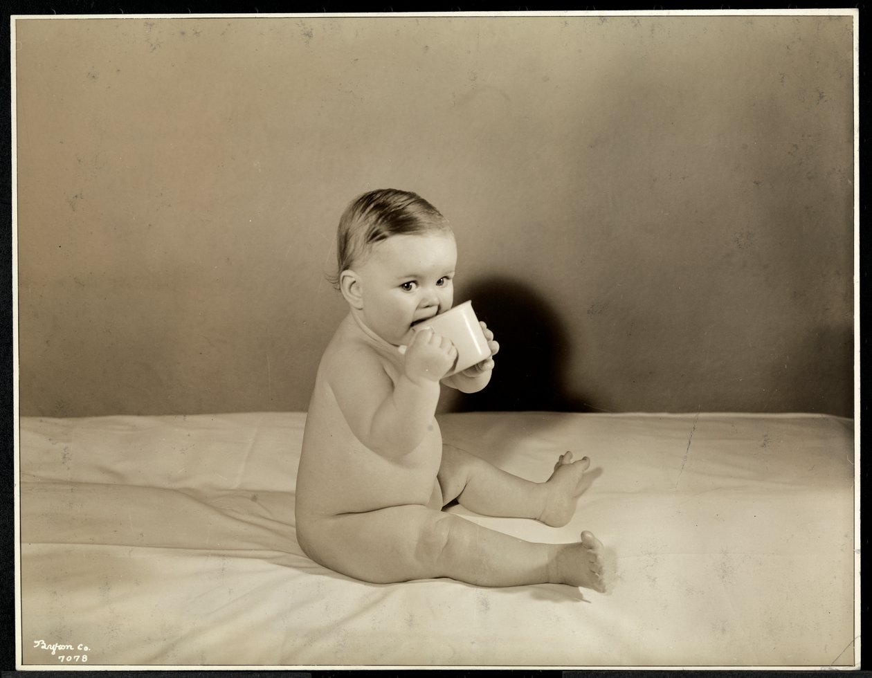 En baby som sitter oppe i sengen og drikker av en kopp på Maternity Center Association i 70th Street East River, New York, 1935 (sølv gelatintrykk) av Byron Company