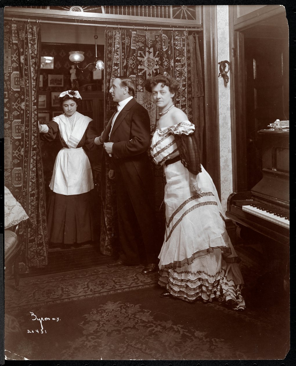 En formelt kledd mann og kvinne og det som ser ut til å være en hushjelp i et rom med piano, 1905 (sølv gelatintrykk) av Byron Company