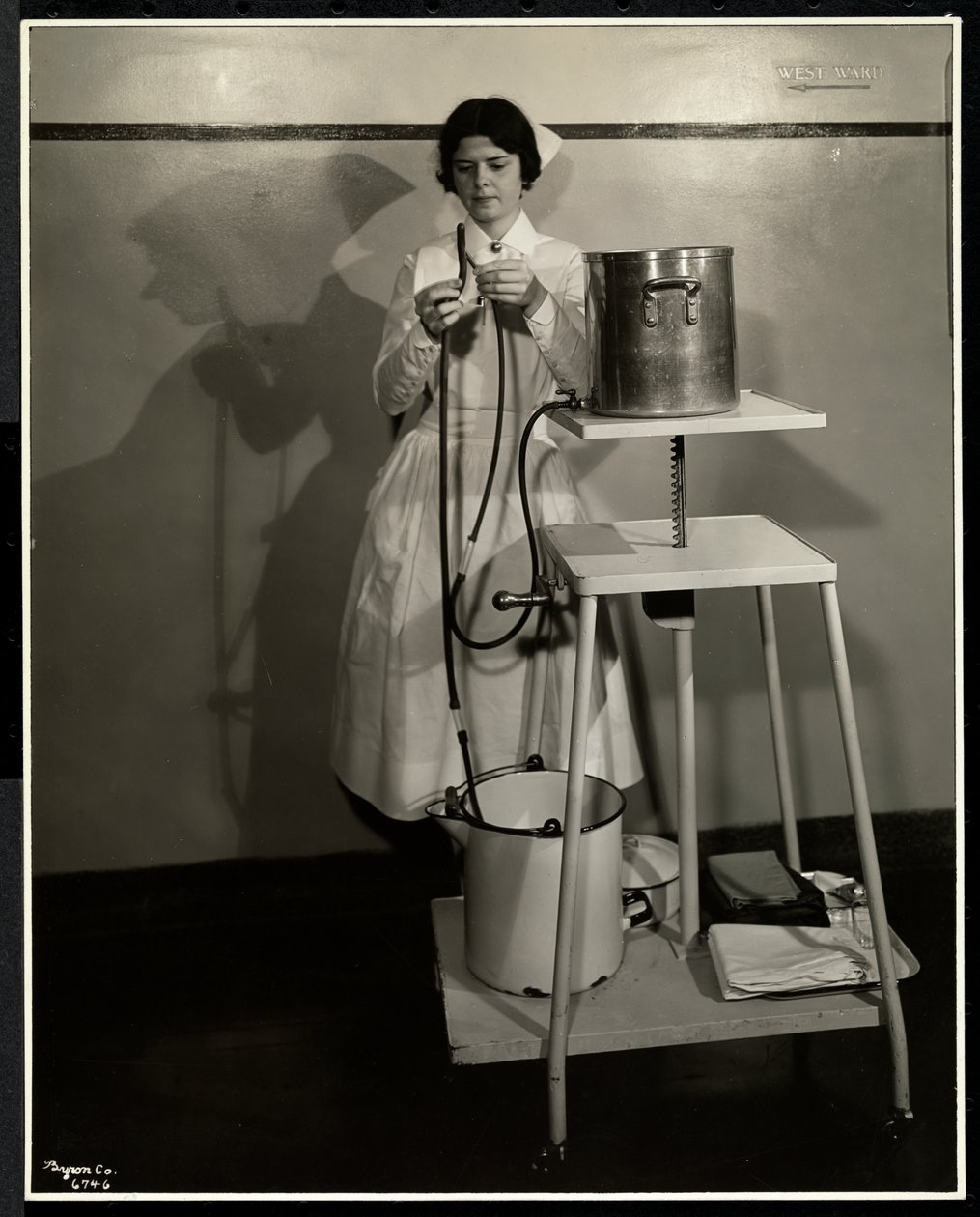 En sykepleier demonstrerer et spesielt apparat med rør og forsynings- og avfallsbøtter ved Sloan Maternity Hospital, Broadway og 168th Street, New York, 1932 (sølv gelatintrykk) av Byron Company