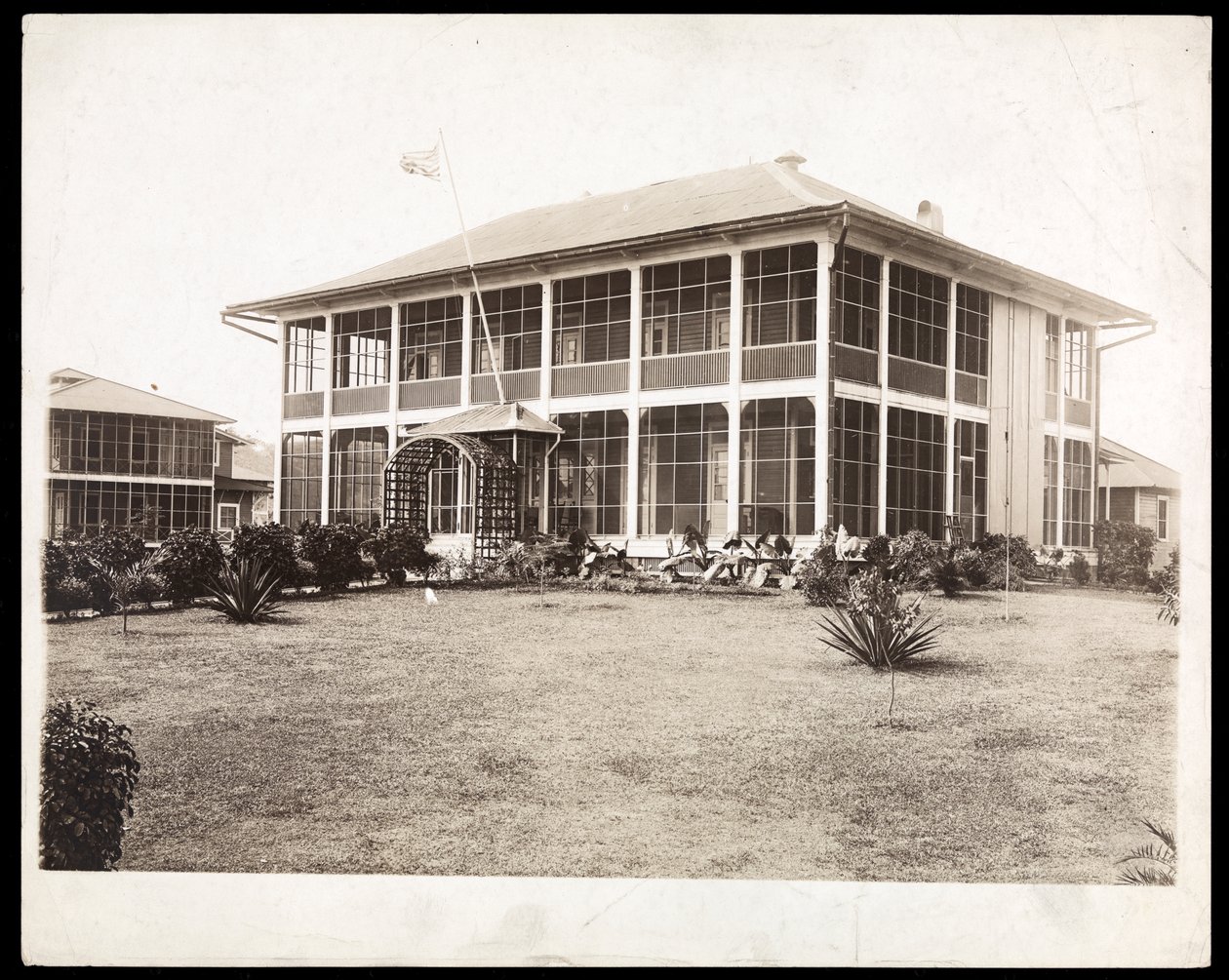 Et to-etasjers hus omgitt av verandaer, med et amerikansk flagg vaiende; i Panama under byggingen av kanalen, 1912 (sølv gelatintrykk) av Byron Company