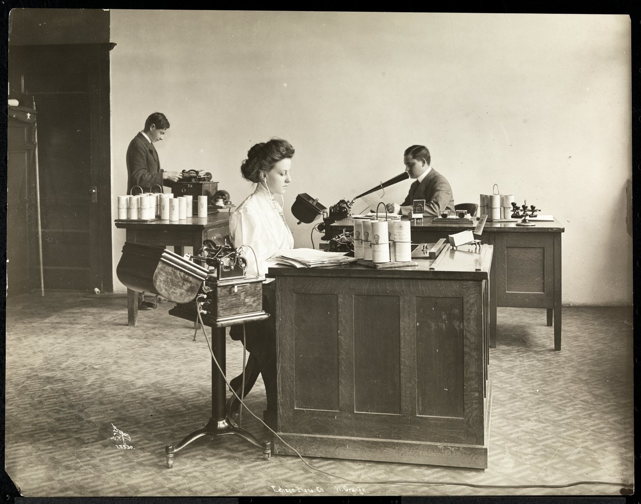 Interiørbilde av en kvinne som skriver fra en diktermaskin og en mann som dikterer inn i en annen ved Edison Phonograph Co., West Orange, New Jersey, 1903 (sølvgelatintrykk) av Byron Company