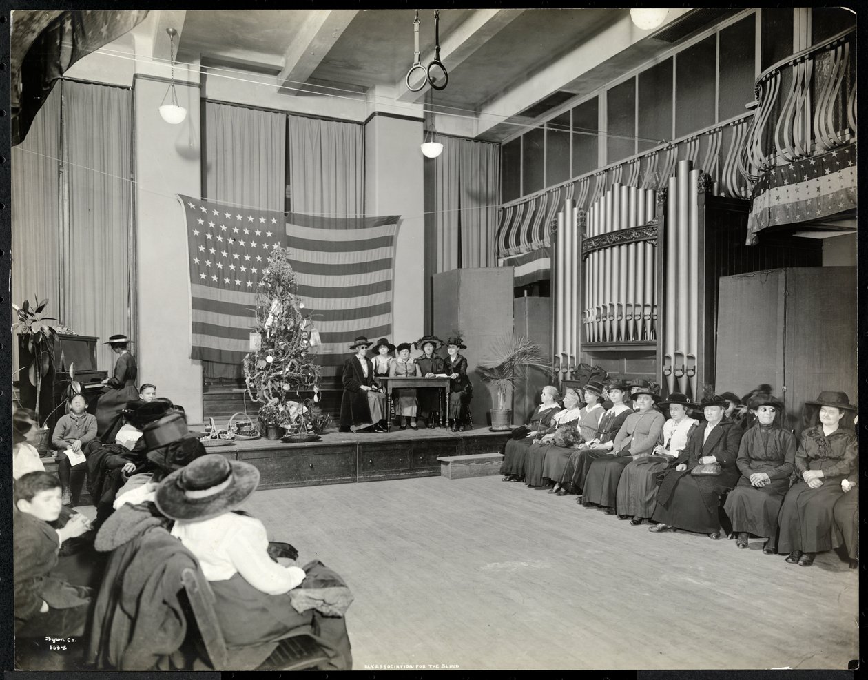 Folk i et auditorium ved New York Association for the Blind med juletre og folk som muligens fremfører en sketsj på scenen med orgel, 1920 (sølv gelatintrykk) av Byron Company