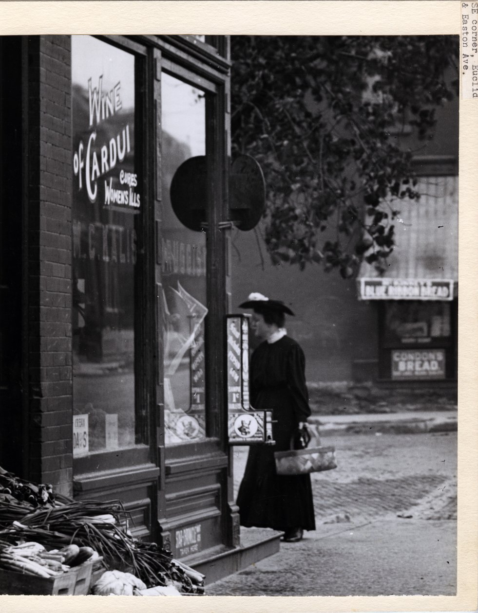 Vertikalt, svart -hvitt fotografi som viser en kvinne som holder en kurv mens hun stod foran inngangen til W. C. Kalis Drugstore på 4878 Easton Avenue av Charles Clement Holt