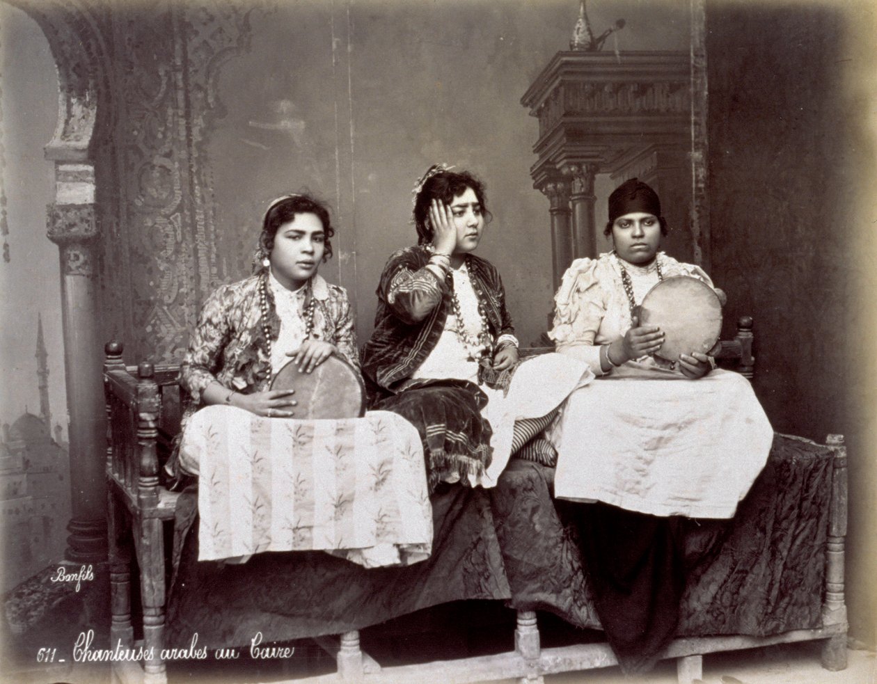 Portrett av tre unge arabiske sangere i etnisk kjole med lange halskjeder. De poserer i studio, sittende. To av dem holder en tamburin av Félix Bonfils