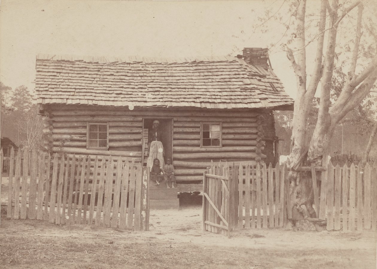 Albumtrykk av en kvinne og to barn foran et tømmerhus i Georgia, 1880-tallet. av H. S. Clark