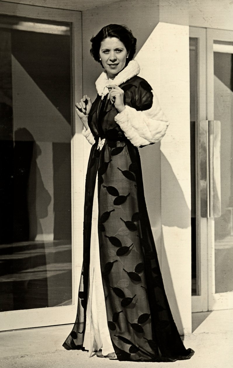 Modellen har på seg en kjole laget av det italienske couture-huset "Bernasconi". Artikkelen som presenteres består av en chiffonfrakk med pelskantet krage og mansjetter (s/hv-bilde) av Luis Bogino