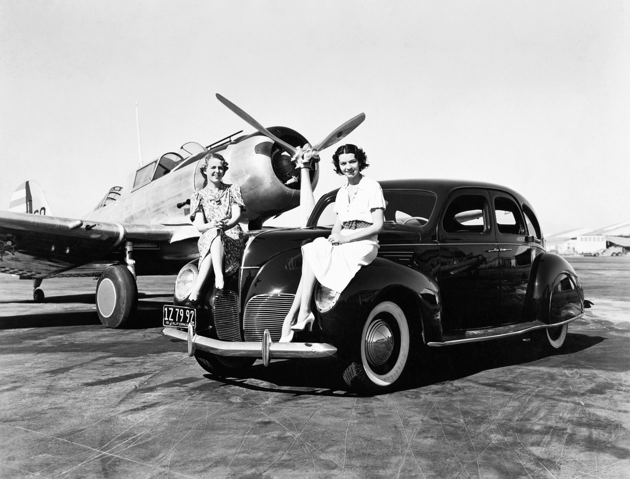 California: 1939 To kvinner sitter på fenderne til en Lincoln Zephyr sedan fra 1939 på en flyplass med et enmotors fly parkert bak seg (s/hv-bilde) av Unbekannt