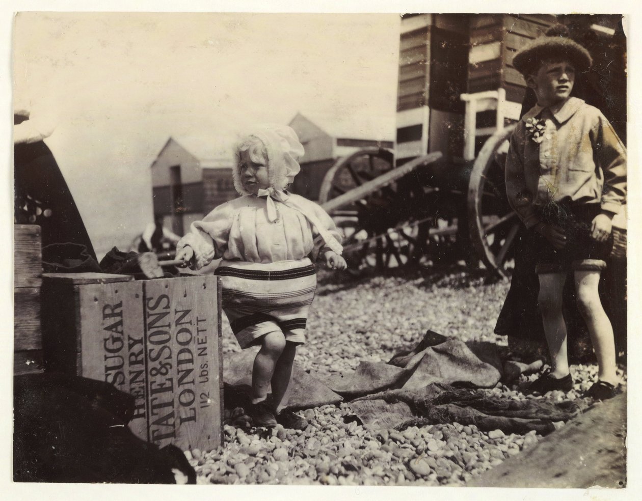 To barn ved bademaskiner, c 1895, Et øyeblikksbilde av to barn ved sjøen, stående blant en gruppe bademaskiner på stranden av Unbekannt