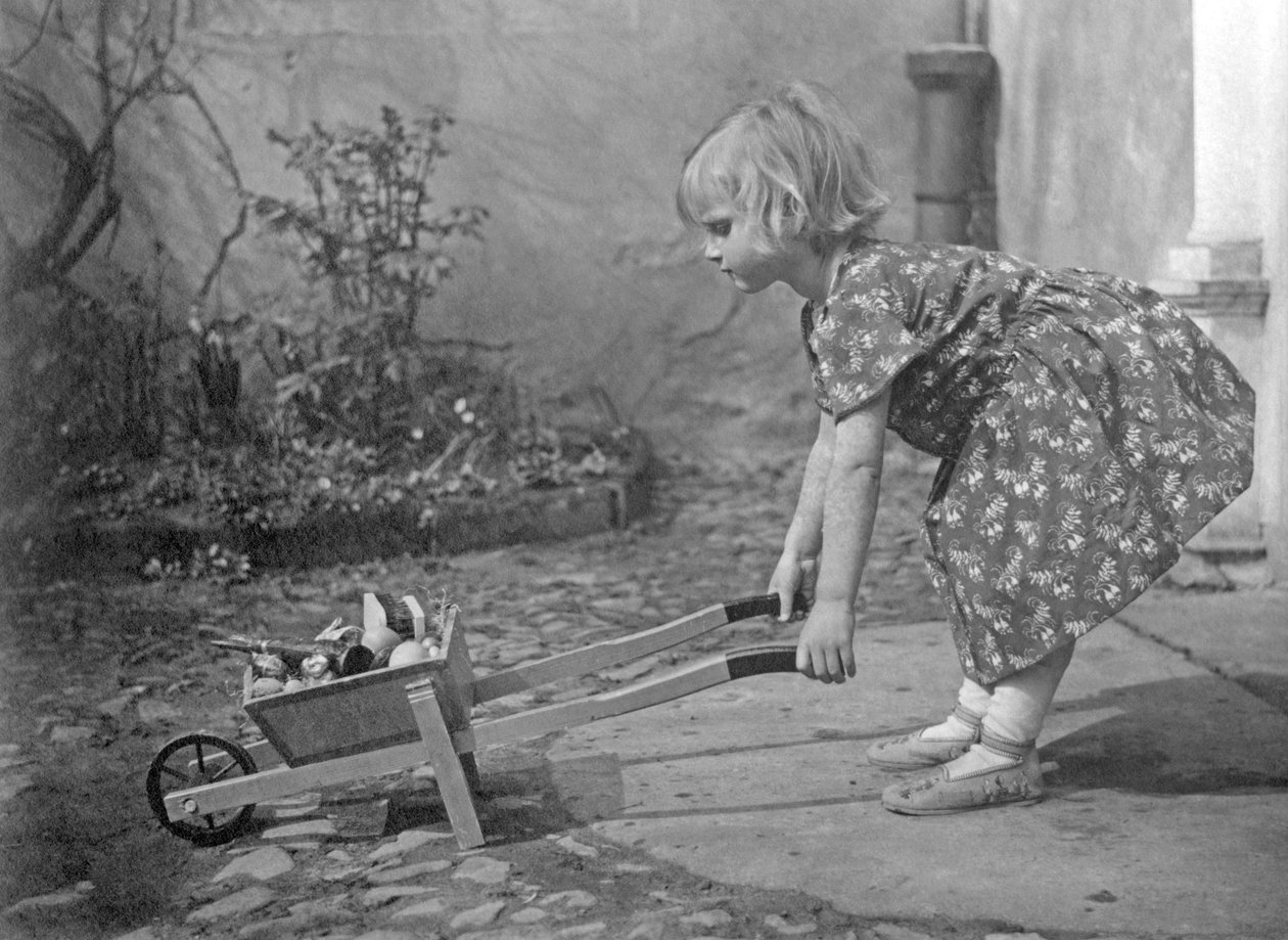 Liten jente leker med en trillebår. Fotografering rundt 1931 av Unbekannt