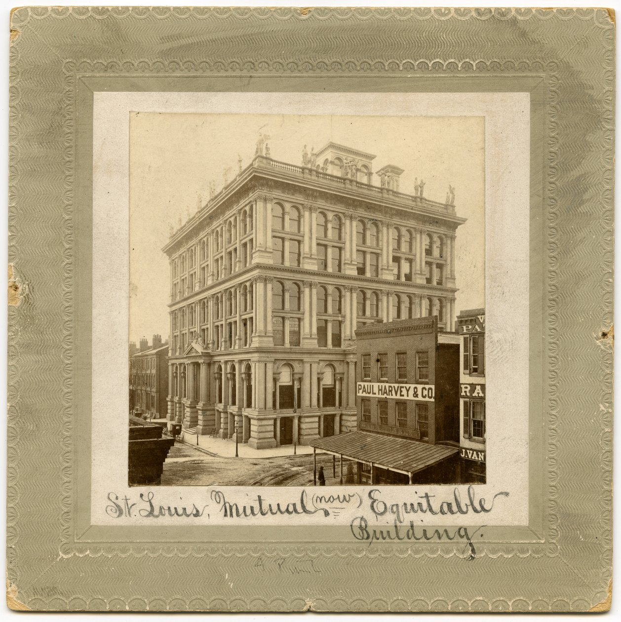 Fotografi av en seks etasjers bygning med figurer langs toppen. Det er andre bygninger synlige på hver side. Bygning også kalt St. Louis Life Insurance Building av Unknown