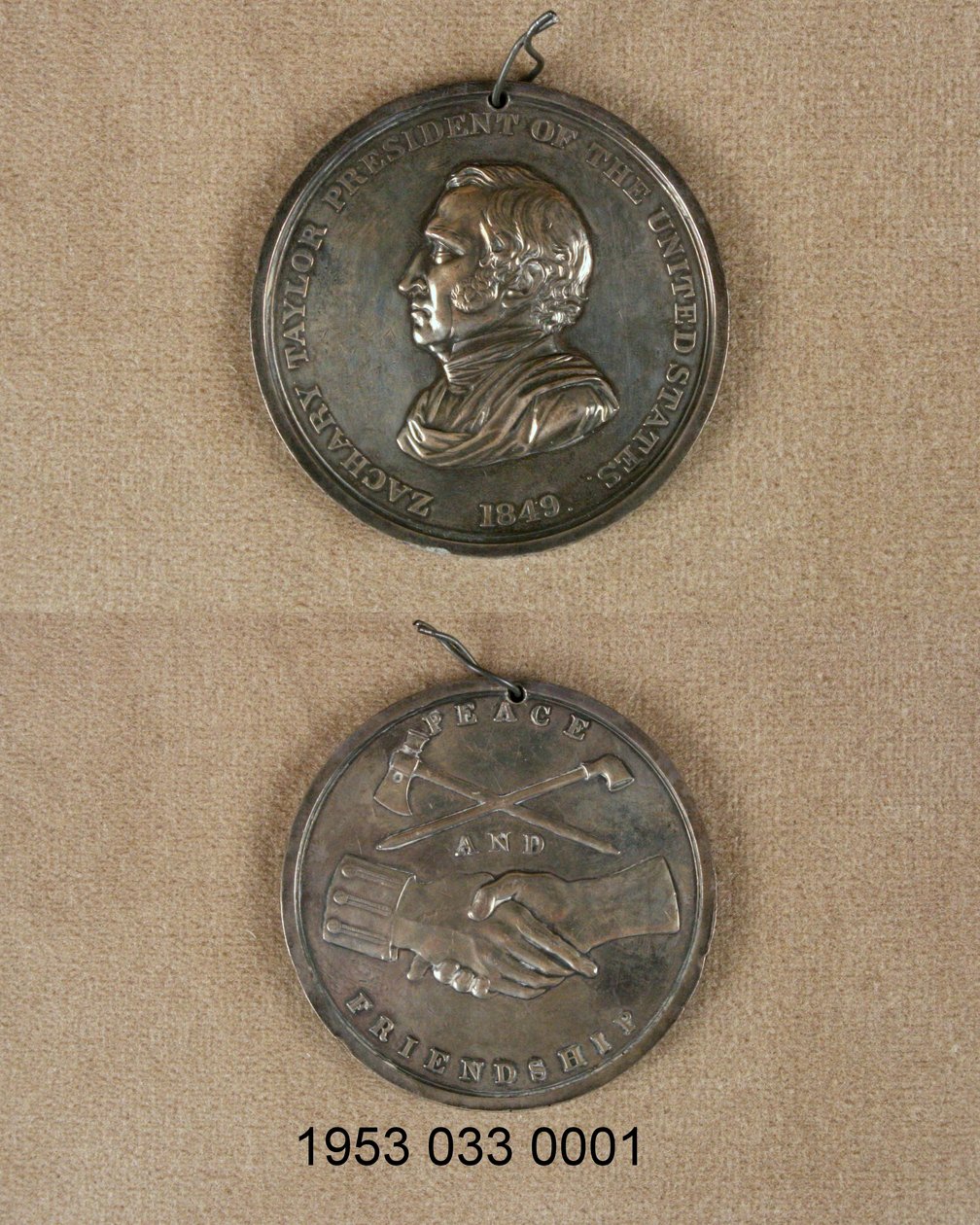 Sølvfredmedalje/forsiden har bysten av Zachary Taylor/baksiden har krysset rør og lem over lukkede hender, en med ensartet mansjett av Unknown