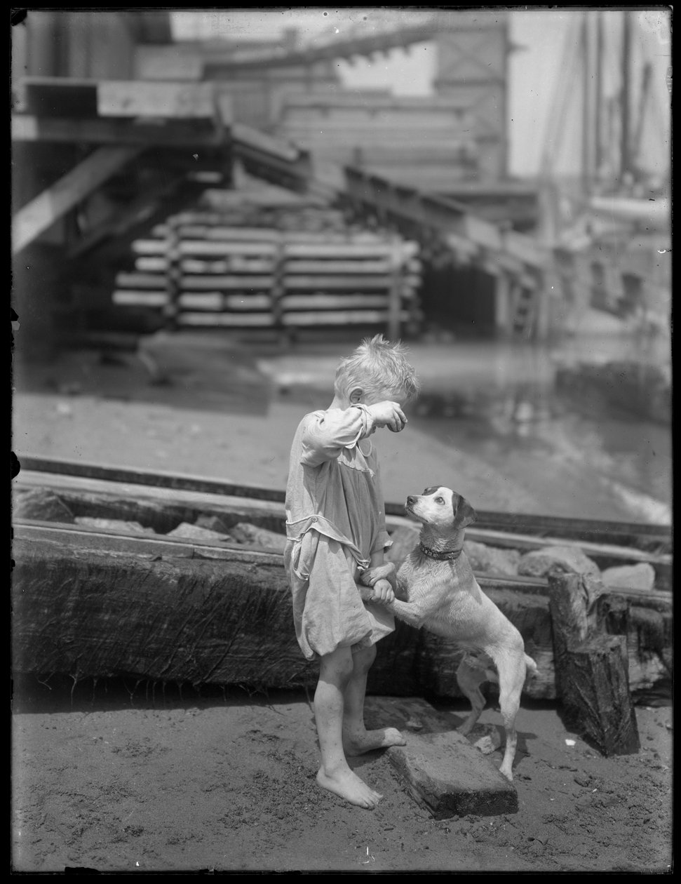 Barfotgutt, muligens William Gray Hassler, leker med hund ved bredden av en elv eller innsjø, New York, ca. 1910 av William Davis Hassler