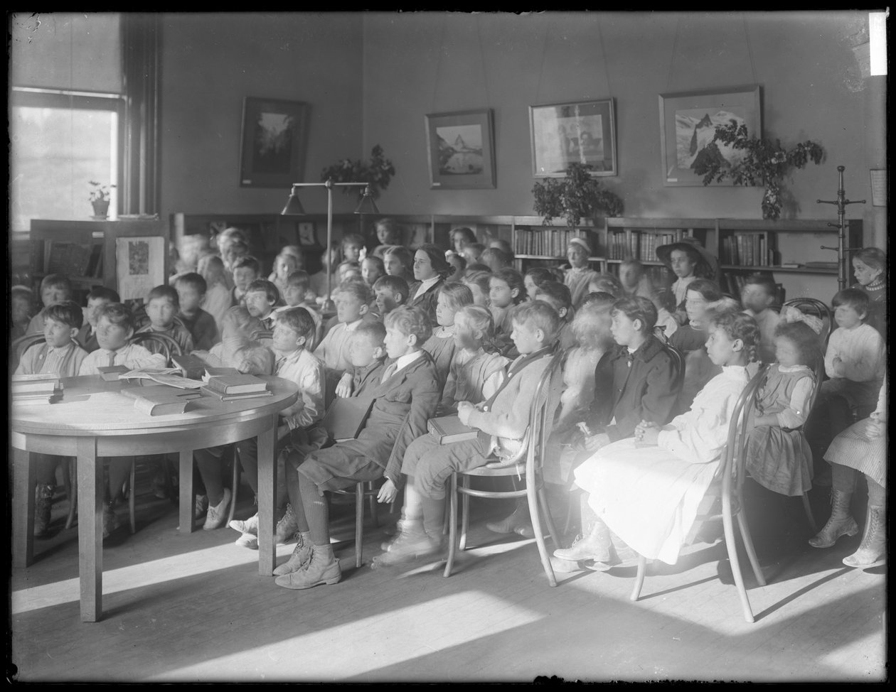 Barn som sitter og lytter på lesesalen til en uidentifisert filial av Queens Borough Public Library, ca. 1910 av William Davis Hassler