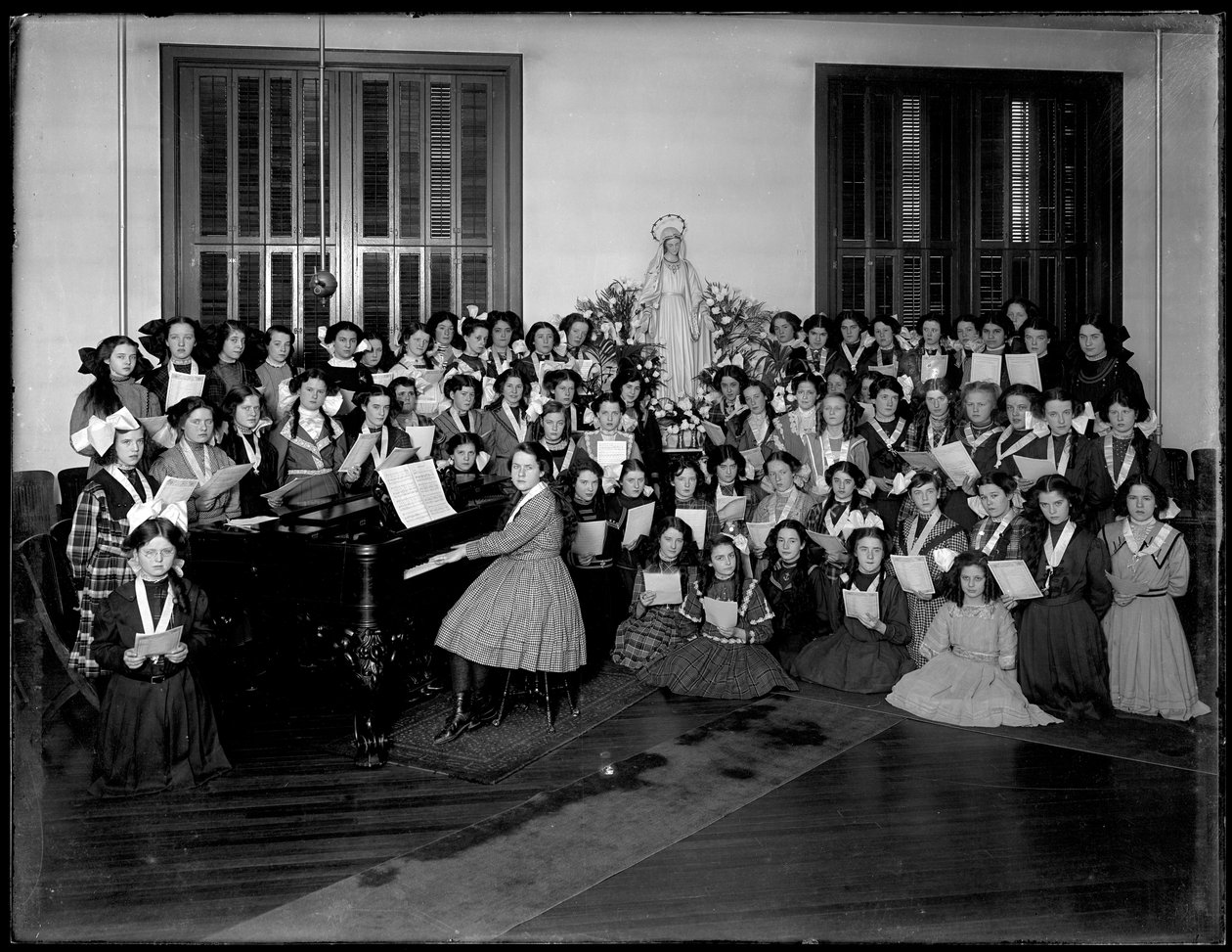 Gruppeportrett av en klasse for jentekor samlet rundt et piano med noter, sannsynligvis på det romersk-katolske orphan asylum i Kingsbridge, Bronx, ca. 1911 av William Davis Hassler