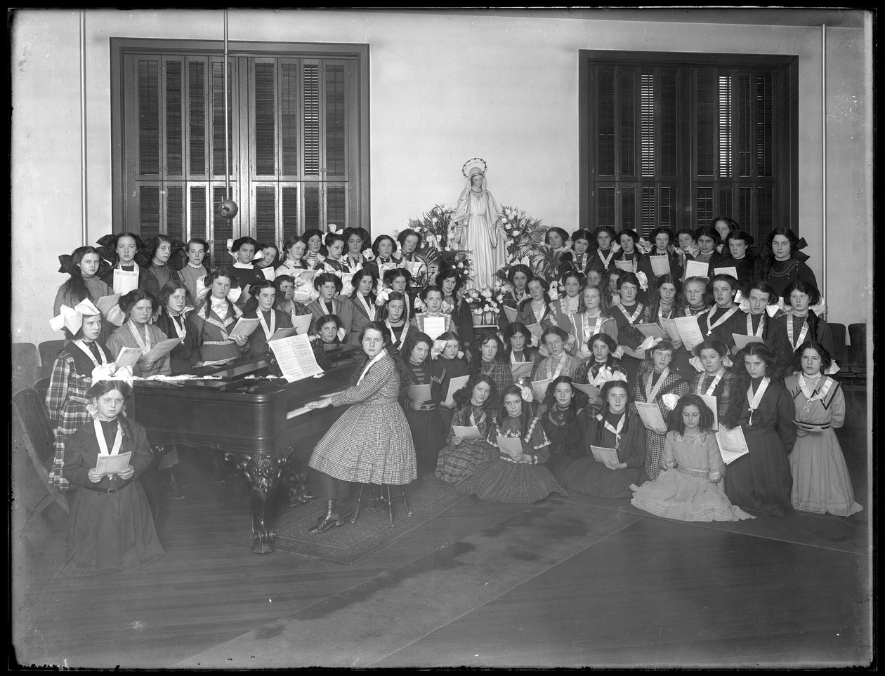 Gruppeportrett av en klasse for jentekor samlet rundt et piano med noter, sannsynligvis på det romersk-katolske orphan asylum i Kingsbridge, Bronx, ca. 1911 av William Davis Hassler