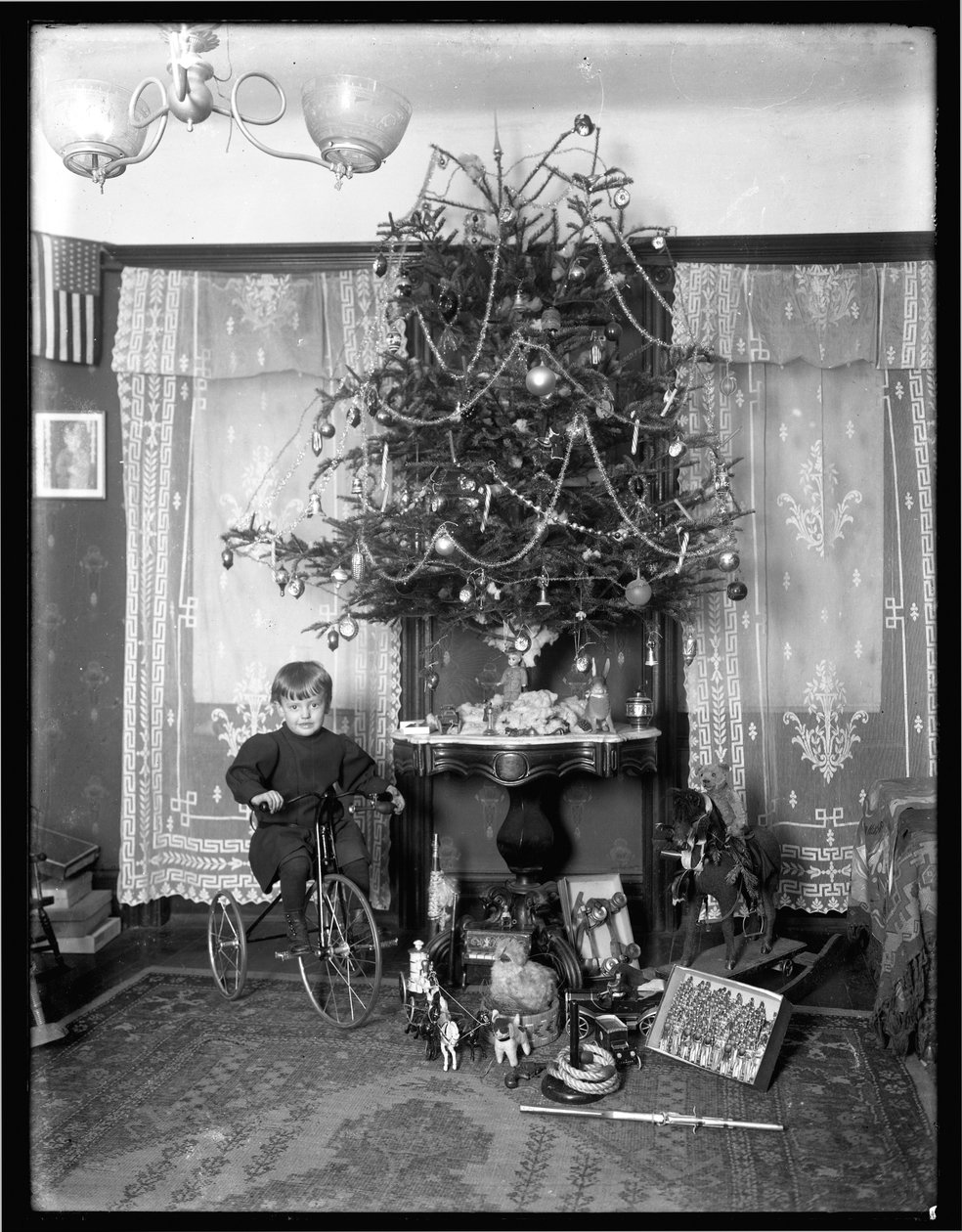 Seymour-gutt poserte sittende på trehjulssykkel ved siden av juletreet i stuen, julen 1912 av William Davis Hassler
