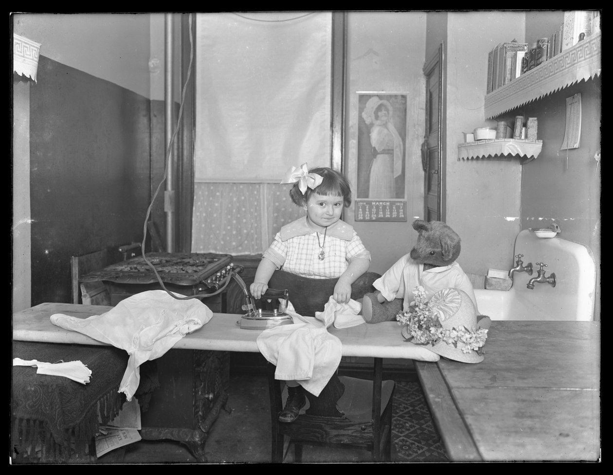 Uidentifisert liten jente som demonstrerer bruken av et elektrisk strykejern, ca. 1913-14 av William Davis Hassler