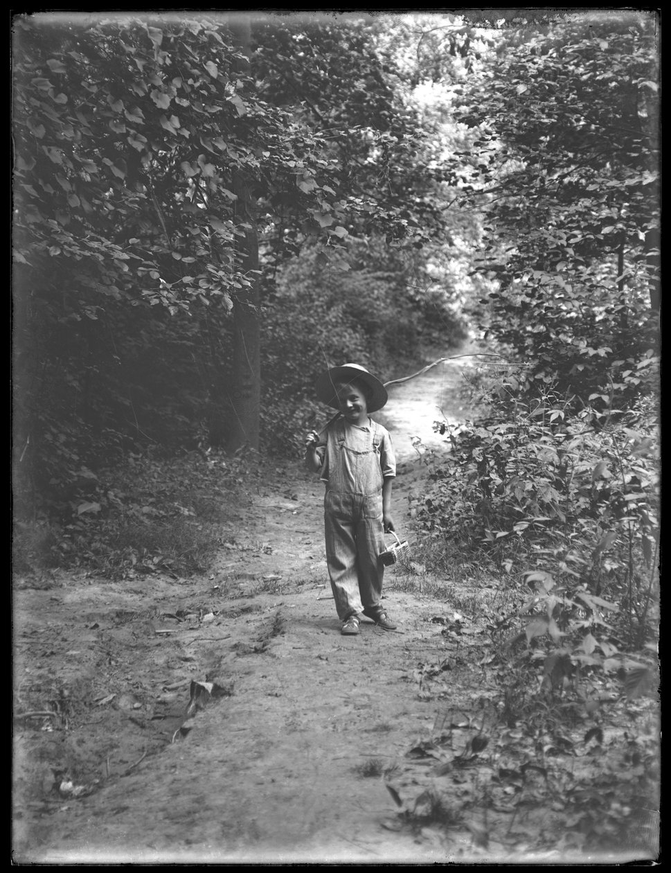 William Gray Hassler i stråhatt og kjeledress, bærende fiskestang og kurv, gående nedover en sti i skogen alene, 20. august 1911 av William Davis Hassler
