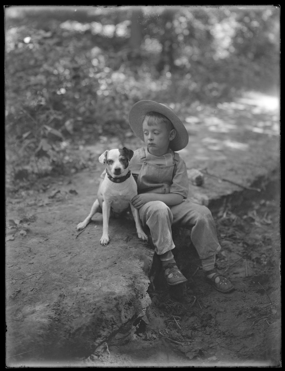 William Gray Hassler i stråhatt og kjeledress, poserte sittende i skogen med en liten hund (Bessie), 20. august 1911 av William Davis Hassler