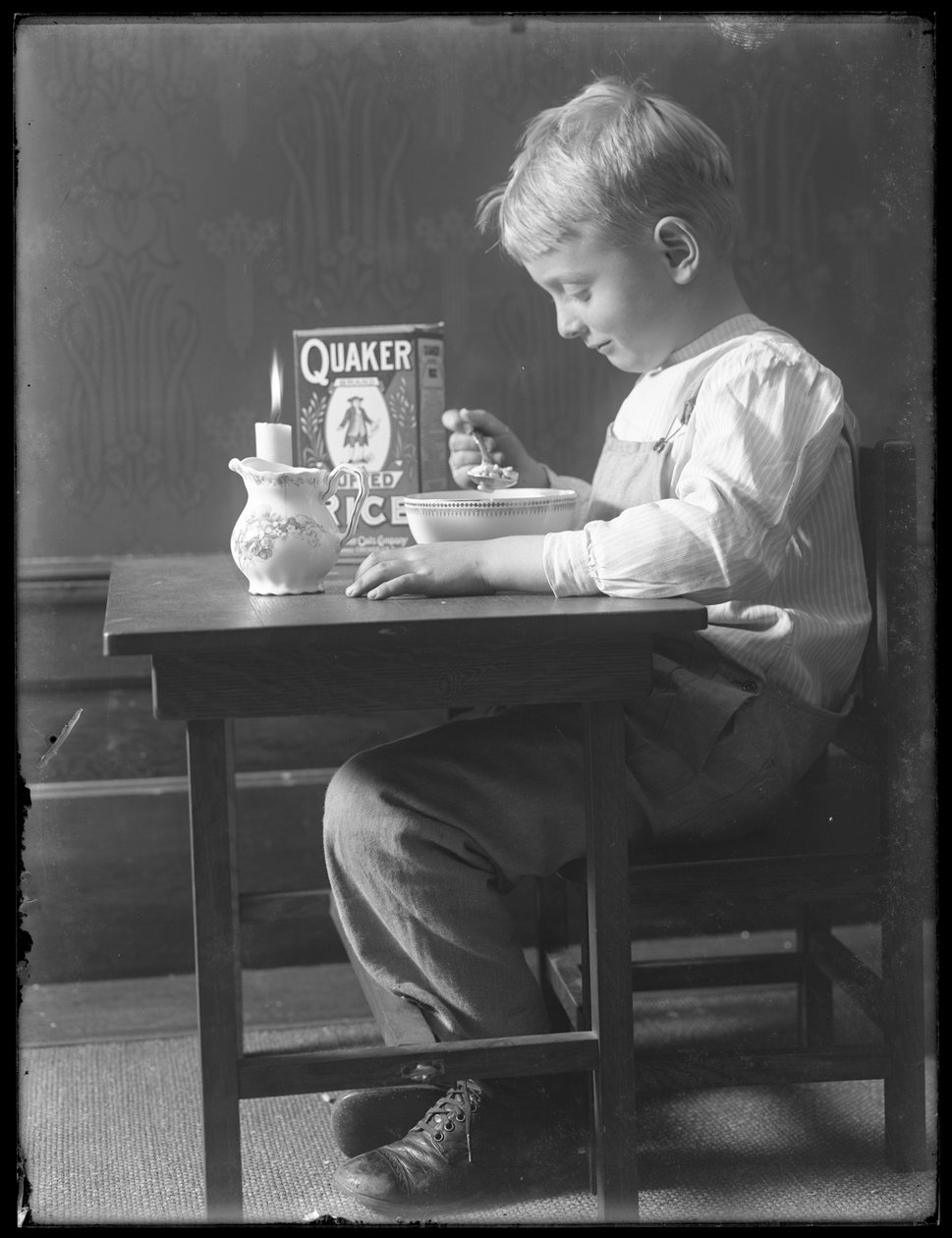William Gray Hassler satt ved et lite bord og spiste en bolle med Quaker-puffet risblanding, ca. 1912 av William Davis Hassler