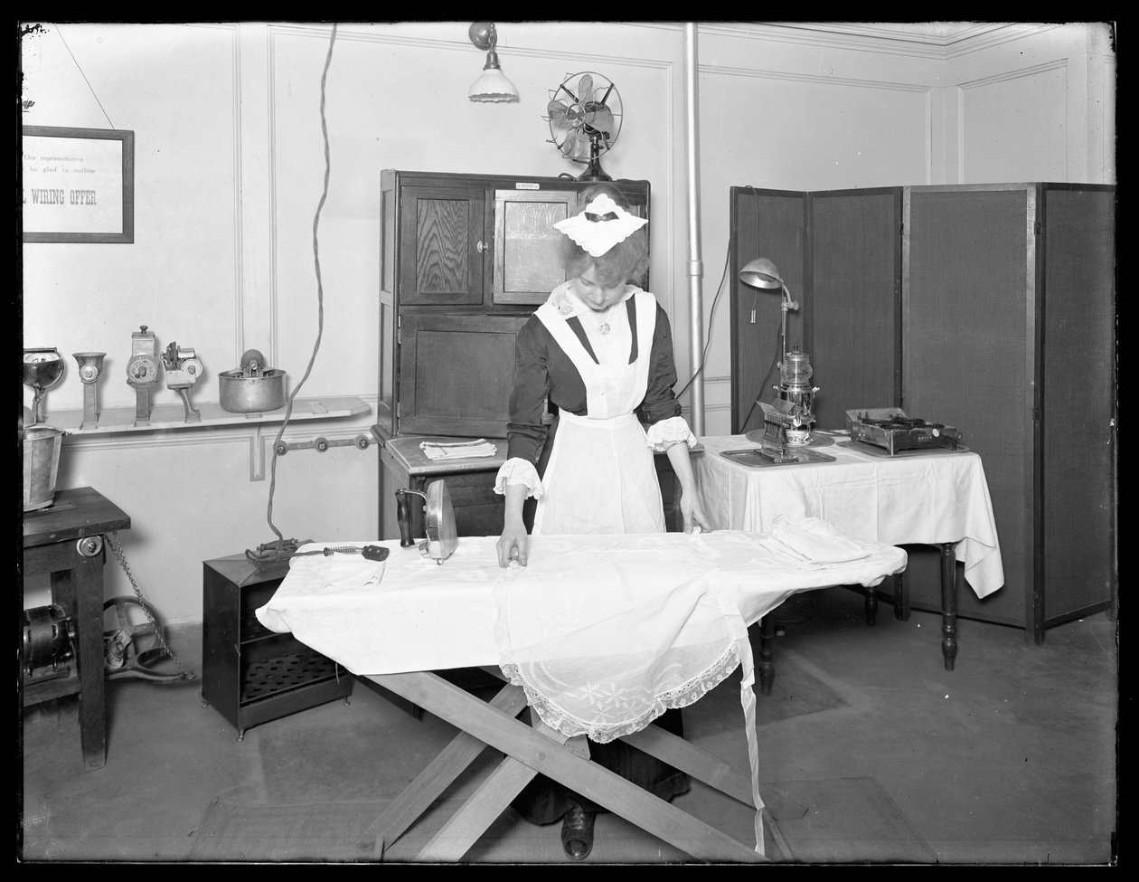 Ung kvinne i hushjelpkostyme som demonstrerer bruken av et elektrisk strykejern, ca. 1913-14 av William Davis Hassler
