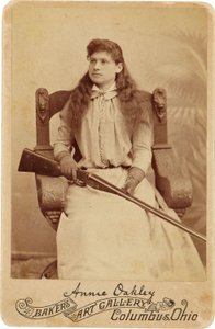 Annie Oakley: Annie Oakley:. Her sitter den legendariske skarpskytteren ganske nedverdig i en massiv stol i renessansestil, en hagle som hviler i fanget hennes. Hennes delikate feminine trekk og flytende hår motvirker hennes robuste utendørs bilde ©Buyenla av American Photographer