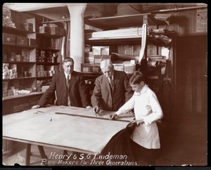 Portrett av Henry og S.G. Lindeman med deres sønn eller barnebarn stående over butikktegninger for et piano, 1910 (sølv gelatintrykk)