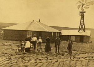 Familie av husmenn, utenfor torvehuset deres med en vindmølle på taket av den tilstøtende bygningen i Custer County, Nebraska, ca. 1885 (s/hvitt-bilde) av Solomon D. Butcher