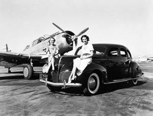 California: 1939 To kvinner sitter på fenderne til en Lincoln Zephyr sedan fra 1939 på en flyplass med et enmotors fly parkert bak seg (s/hv-bilde) av Unbekannt