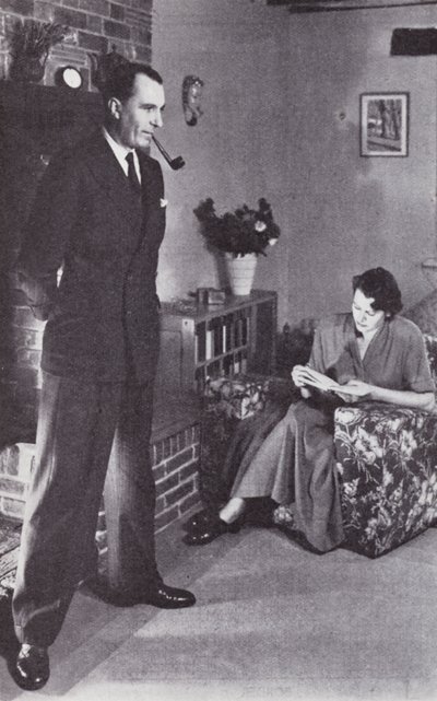 Kona leser, mens mannen varmer ryggen mot bålet, ca.1950 av English Photographer