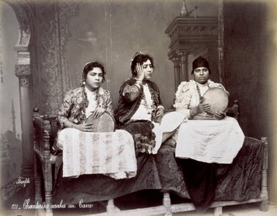 Portrett av tre unge arabiske sangere i etnisk kjole med lange halskjeder. De poserer i studio, sittende. To av dem holder en tamburin av Félix Bonfils