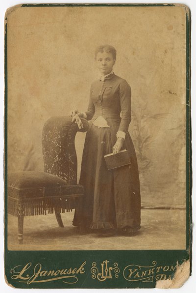 Fotografi av en kvinne som står ved siden av en stol og holder en bok, sent på 1800-tallet. av Louis Janousek