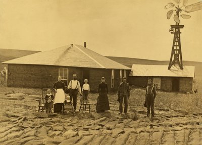 Familie av husmenn, utenfor torvehuset deres med en vindmølle på taket av den tilstøtende bygningen i Custer County, Nebraska, ca. 1885 (s/hvitt-bilde) av Solomon D. Butcher