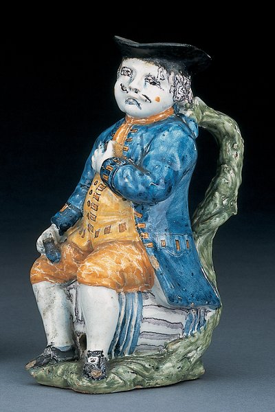 Tobymugge med ansikt og hender i blekt mangan, holdende og tom kulebrunfarget kanne i vasket blå jakke og knebukser, og grønn vest, ca. 1800 av Unbekannt