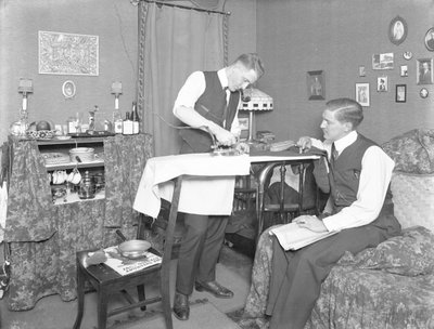 Uidentifisert mann som demonstrerer bruken av et elektrisk strykejern, overvåket av en annen mann, ca. 1913-14 av William Davis Hassler