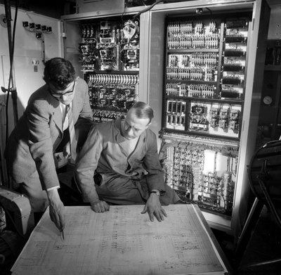 Ingeniører diskuterer planer foran det elektroniske kontrollpanelet de har bygget av Frederick (attr. to) Barnard