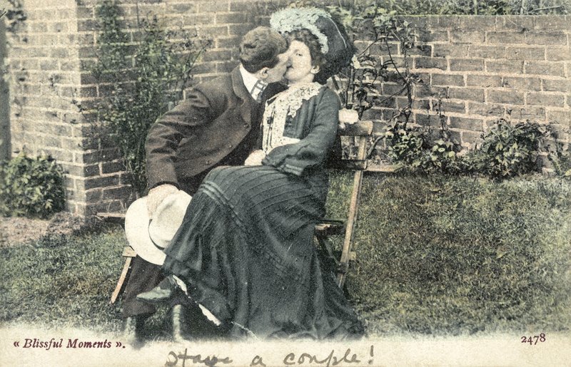 Mann som tar et kyss fra en jente på en hagebenk (fargebilde) av English Photographer
