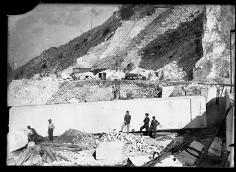 Tre menn observerer arbeidet til flere gravemaskiner i Apuan-alpene. Bak dem er en enorm blokk med marmor som allerede er firkantet og klar til å bli transportert til dalen (s/hvitt-bilde)