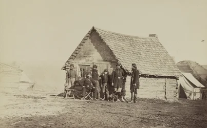 En gruppe soldater og to unge menn, en afroamerikaner, står utenfor tømmerhyttekvarteret, 1861-65