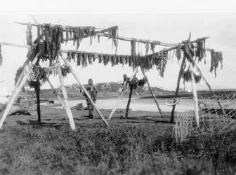En eskimomann som står under en struktur med hvalkjøtt hengende til tørk, ca. 1929 (s/hvitt-bilde)