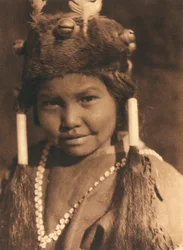 Nærbilde av en amerikansk indianergutt fra Klamath-stammen eller -landsbyen. Han har en pelshatt med påsatte eikenøtter og fjær og håret i to hestehaler