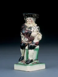 Dick Whittington kanne, støpt opp fra en stol med høy rygg, holder katten sin, sprutet i brune, grønne og svarte klær, et støpt skjold ved siden, ca. 1790