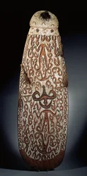 Ovalt Asmat-skjold, skåret i relieff med rullende ornament som inneholder svært stiliserte figurer, flettede dusker til kanten (tre, raffia og fjær)