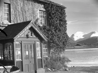 Utsikt over en inngang til «Anglers Hotel», innsjø og fjell til høyre for bildet