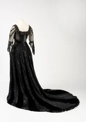 Svart ankellang aftenkjole båret av dronning Alexandra, med perle- og paljettdekorasjon og med dronning Alexandras karakteristiske svarte silkebånd med navn i gullbokstaver, laget av Barolet of Knightsbridge, London, 1908-1910 (silke og fløyel)