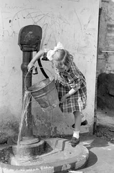 Liten jente tømmer en bøtte med vann i en pumpefontene, 9. mai 1939 (s/h-bilde)