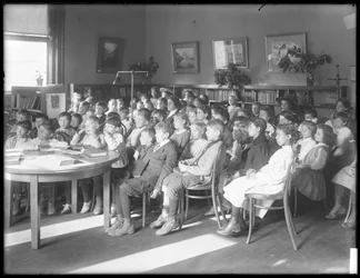 Barn som sitter og lytter på lesesalen til en uidentifisert filial av Queens Borough Public Library, ca. 1910