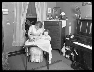 Uidentifisert kvinne demonstrerer bruken av et elektrisk strykejern, overvåket av en liten jente, ca. 1913-14